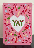 A pink greeting card with a white scalloped heart in the center and the word 'YAY' in green and red. The card is decorated with colorful confetti, stars, and streamers on a pink background.