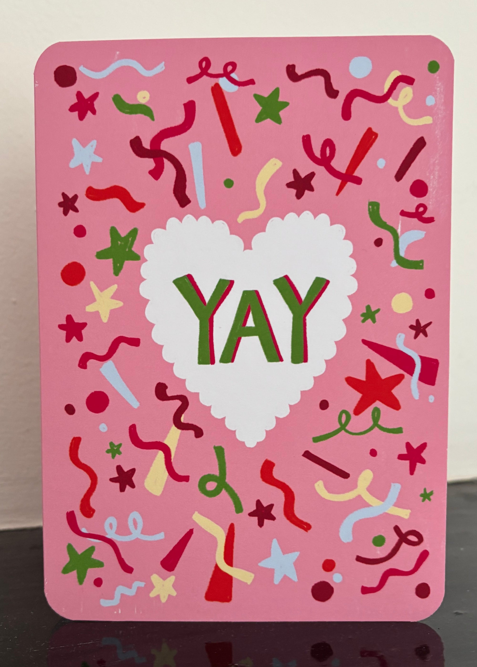 A pink greeting card with a white scalloped heart in the center and the word 'YAY' in green and red. The card is decorated with colorful confetti, stars, and streamers on a pink background.