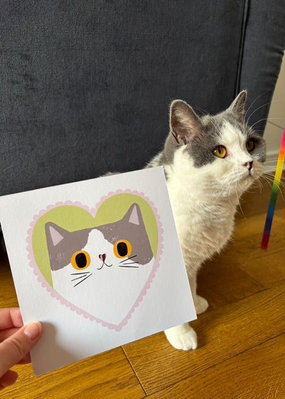 A hand holds a square card featuring a cute illustrated cat face inside a heart, while a real cat sits on a wooden floor in the background near a dark gray upholstered chair.