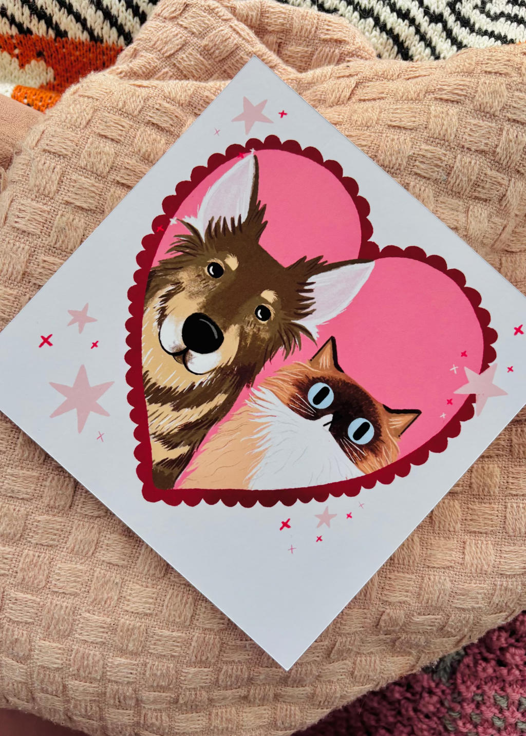 A square custom print featuring a cartoon dog and cat inside a pink heart, placed on a textured beige blanket.