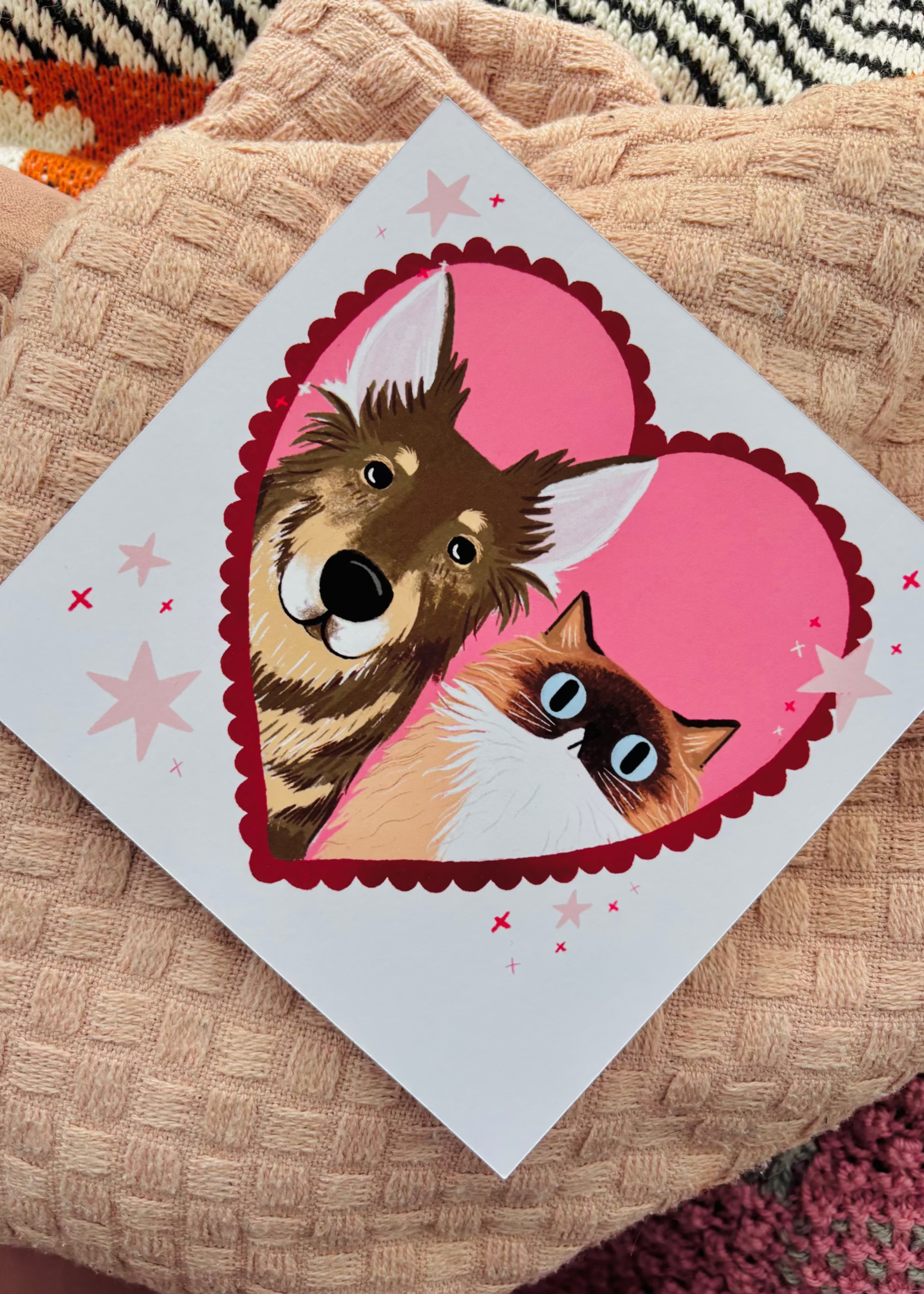 A square custom print featuring a cartoon dog and cat inside a pink heart, placed on a textured beige blanket.