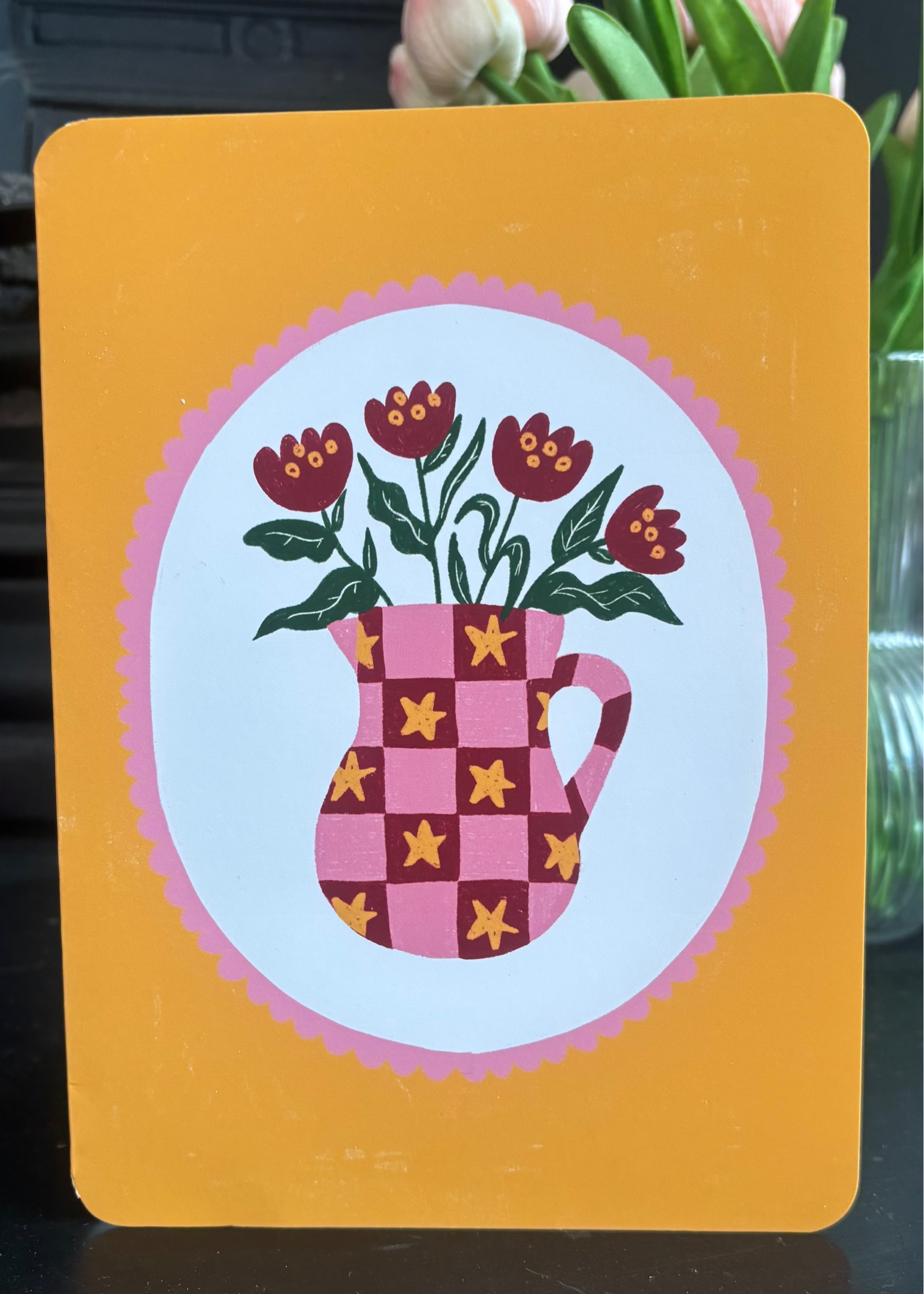A decorative card or print with rounded corners, featuring a yellow-orange border and a central illustration of a pink checkerboard pitcher filled with dark red flowers and green leaves, set against a softly blurred indoor background with flowers.