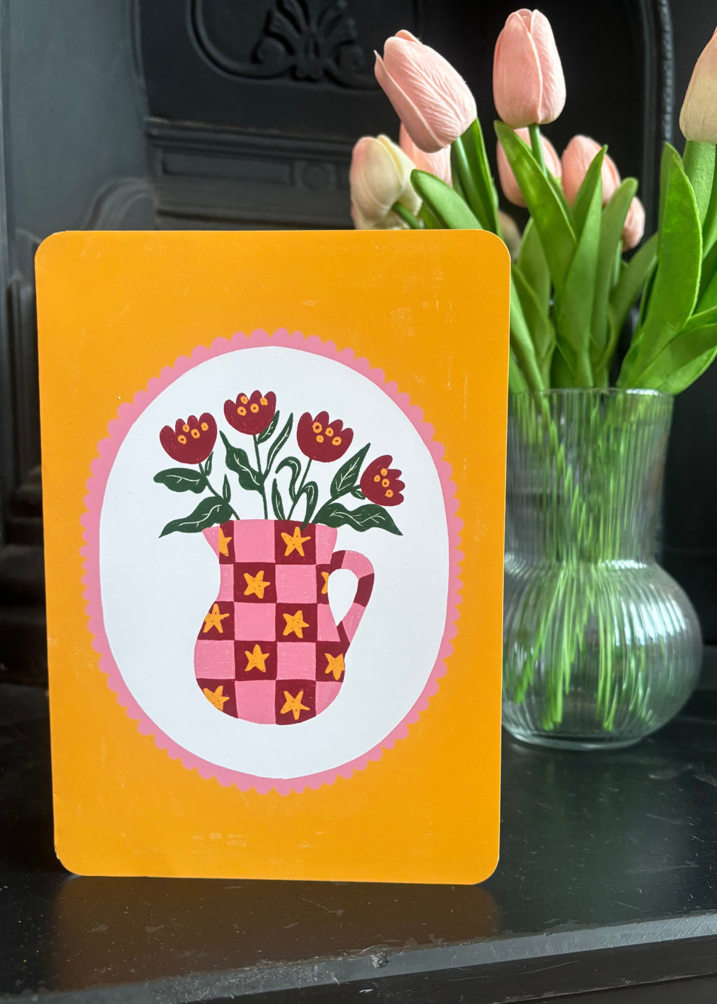 A yellow card or print with a circular white center featuring a pink patterned mug and red flowers, placed on a dark surface next to a glass vase with pink tulips.