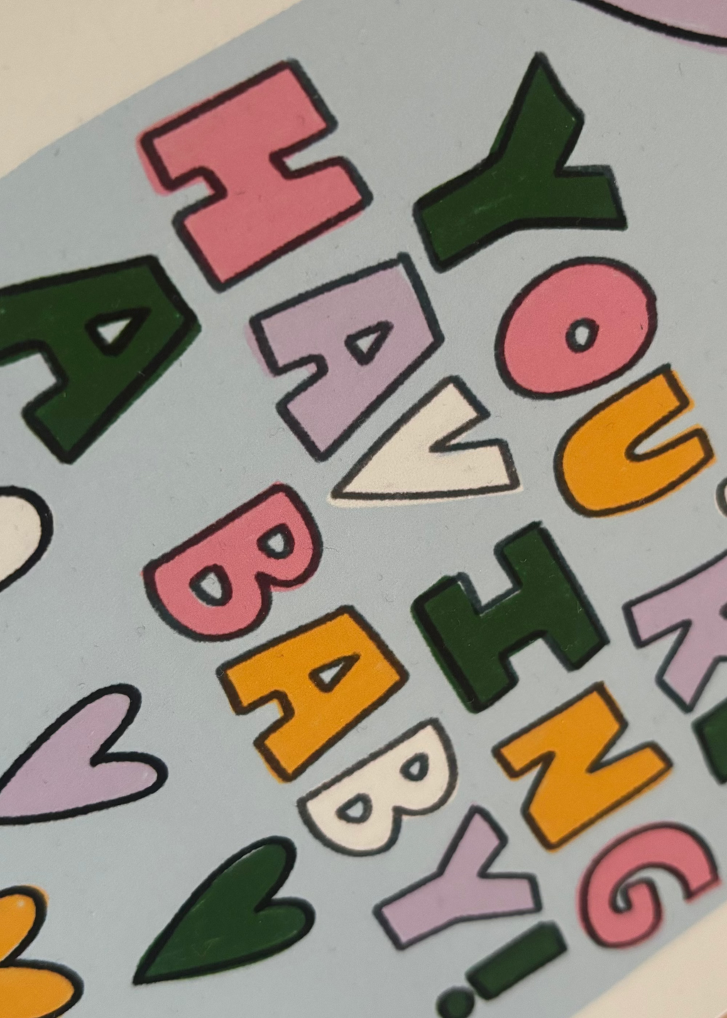 A close-up of a  card with rounded corners featuring the text 'Youre having a baby!' in colorful retro letters and pink, purple, green, yellow, and white hearts on a light blue background.