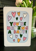 A greeting card with a white border and a light blue center panel that reads “YOU’RE HAVING A BABY!” in colorful playful letters, surrounded by pastel hearts. The card is photographed on a dark tabletop with pink tulips in a glass vase behind it.