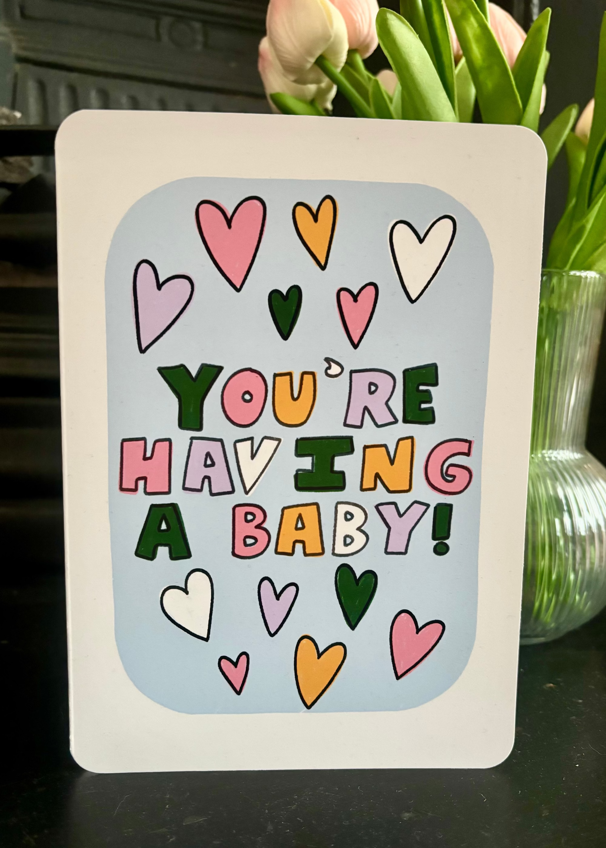 A greeting card with a white border and a light blue center panel that reads “YOU’RE HAVING A BABY!” in colorful playful letters, surrounded by pastel hearts. The card is photographed on a dark tabletop with pink tulips in a glass vase behind it.