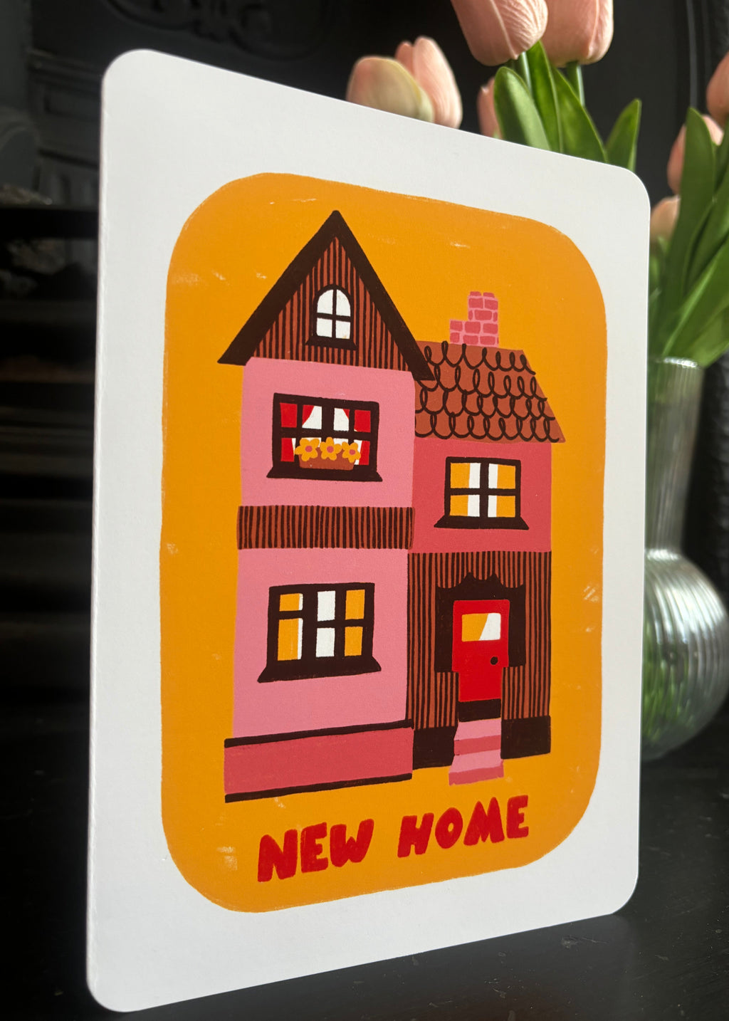 A square white card or print featuring a stylized pink house illustration on an orange background with the words 'NEW HOME' in red at the bottom, photographed against a dark indoor background with blurred flowers.