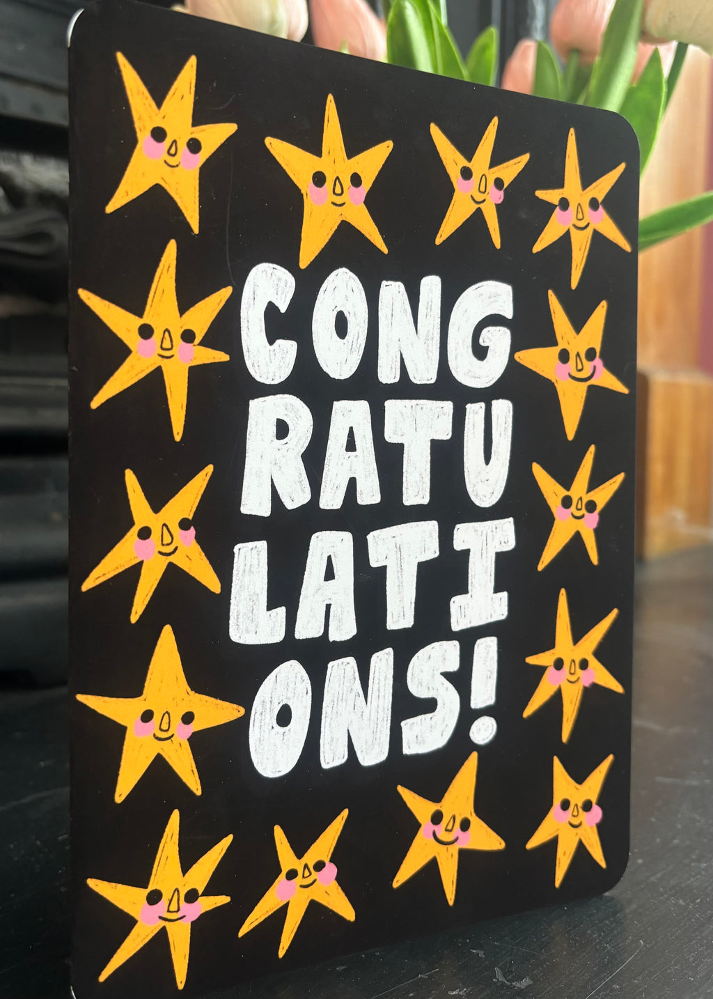 A black rectangular sign with rounded corners featuring the text 'CONGRATULATIONS!' in large white letters, surrounded by yellow smiling star illustrations with pink cheeks, photographed indoors on a dark surface with blurred background elements.