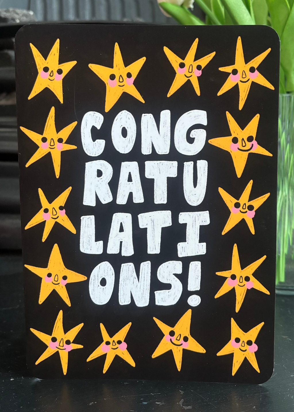 A rectangular black sign with the words 'CONGRATULATIONS!' in large white letters, surrounded by yellow smiling star illustrations, photographed on a tabletop with flowers and furniture in the background.