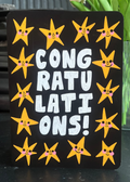 A rectangular black sign with the words 'CONGRATULATIONS!' in large white letters, surrounded by yellow smiling star illustrations, photographed on a tabletop with flowers and furniture in the background.