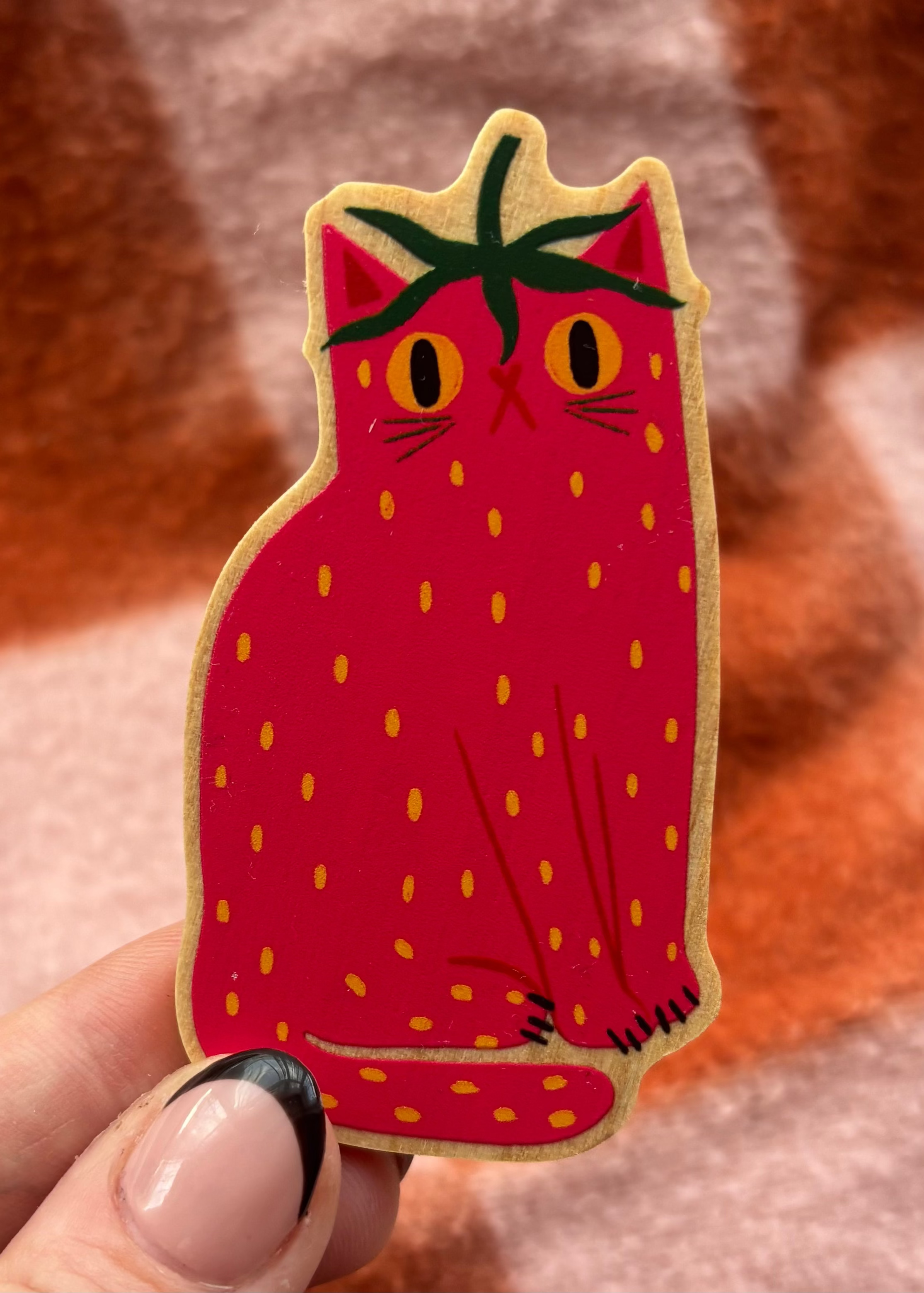 A strawberry-shaped cat pin with a red body, yellow seeds, green leaves on top, and a beige outline, held by a hand against a soft brown and pink background.