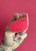 A hand  holds a small strawberry loafing cat with pink skin and yellow eyes and strawberry seeds against a solid pink background.