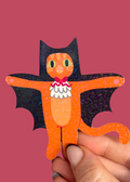 A hand holding a small orange bat-shaped craft figure with black wings, yellow eyes, and a white ruffled collar against a solid pink background.